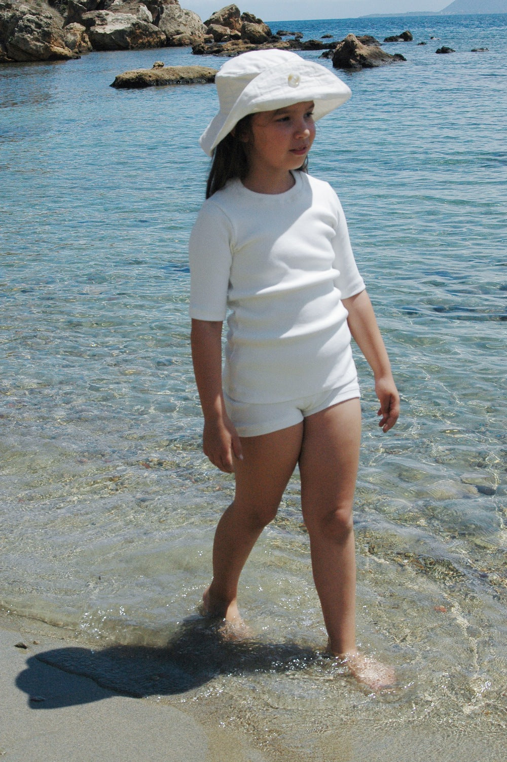 A young girl dressed in the CLASSIC SET baby + kid from cabane childrenswear, complete with a white sunhat, stands at the edge of the sea as gentle waves lap at her feet. She gazes out, enjoying the sunny day. Rocky formations serve as a backdrop, while the water remains clear and blue.