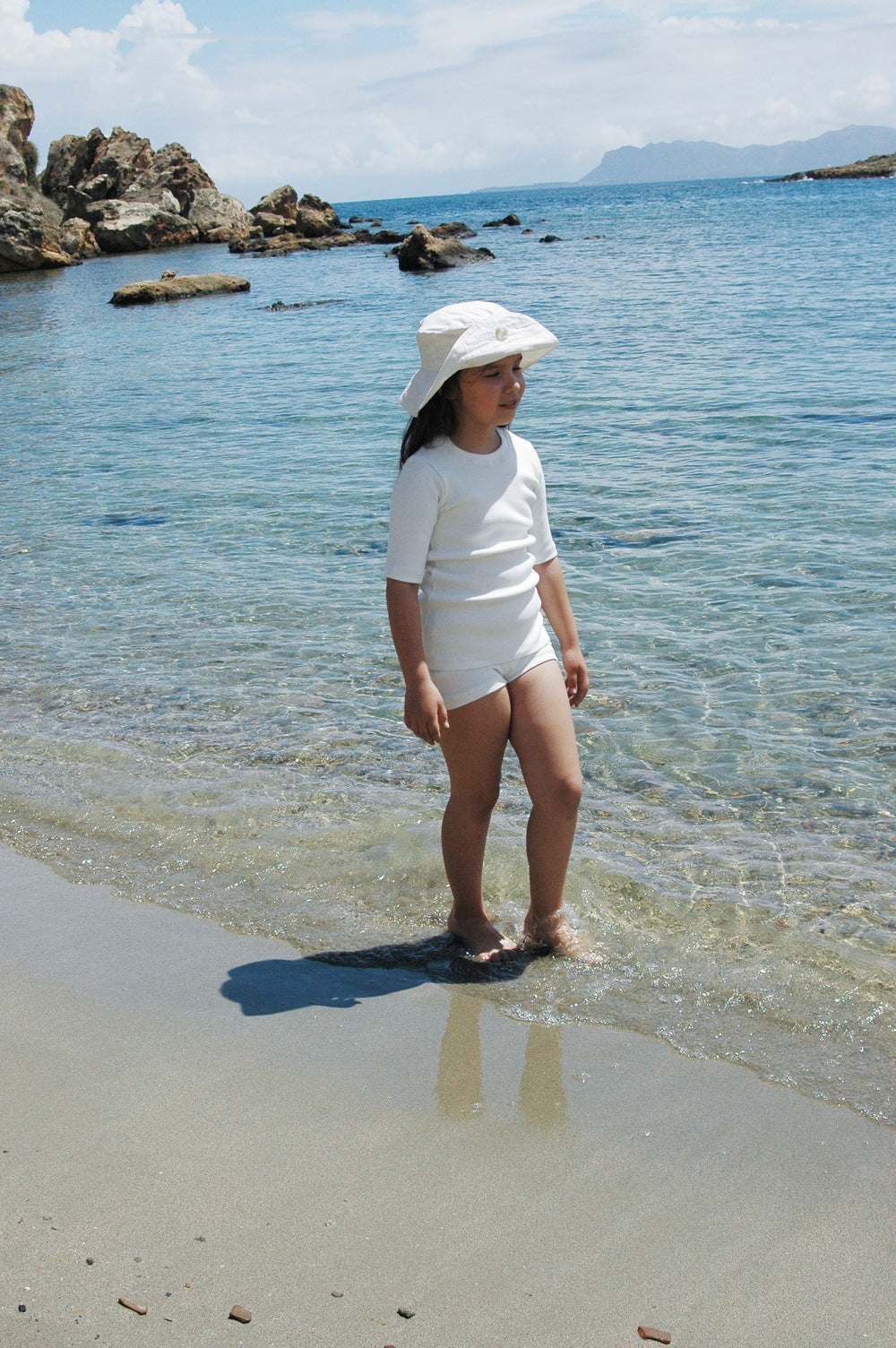 A child stands at the edge of the shore, with clear blue water and rocky formations in the background. Clad in a white CLASSIC SET from cabane childrenswear featuring rib texture and a wide-brimmed hat, the child enjoys a sunny day at the beach. Their attire is part of an ethically handmade collection that complements the serene scene perfectly.