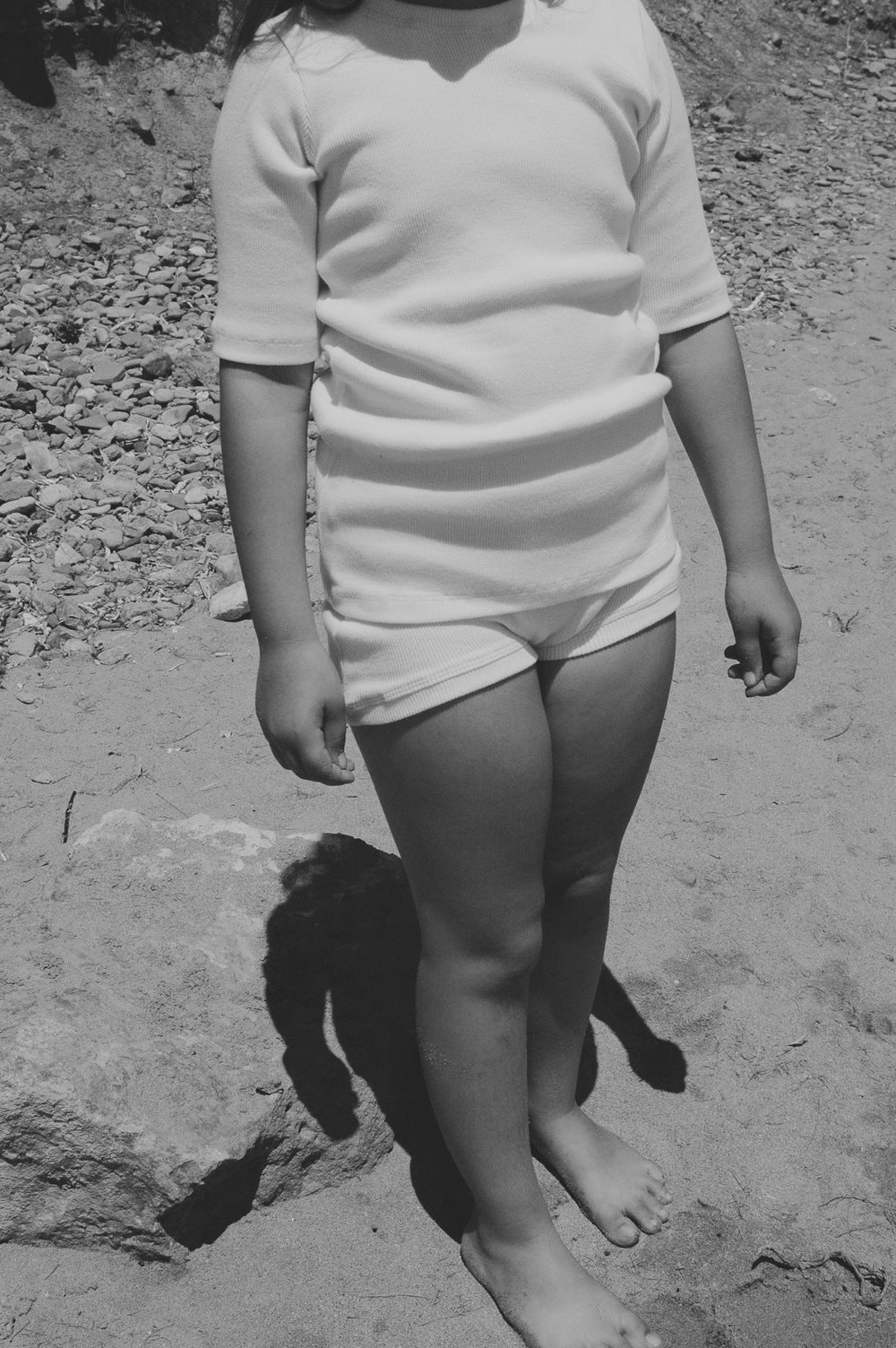 A black-and-white photo of a young child standing outdoors on a sandy surface, with their upper body cropped out of the frame. The child is wearing an ethically handmade CLASSIC SET baby + kid from cabane childrenswear, consisting of a light-colored short-sleeve shirt and matching shorts. Shadows of their legs are visible on the ground, showcasing the rib texture fabric.