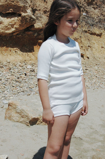 A young child with long hair stands on a sandy beach near some rocky terrain, wearing the CLASSIC SET baby + kid outfit from cabane childrenswear. The ensemble features a white rib-textured long-sleeved top and shorts. The child gazes thoughtfully into the distance, under a sunny and clear sky.