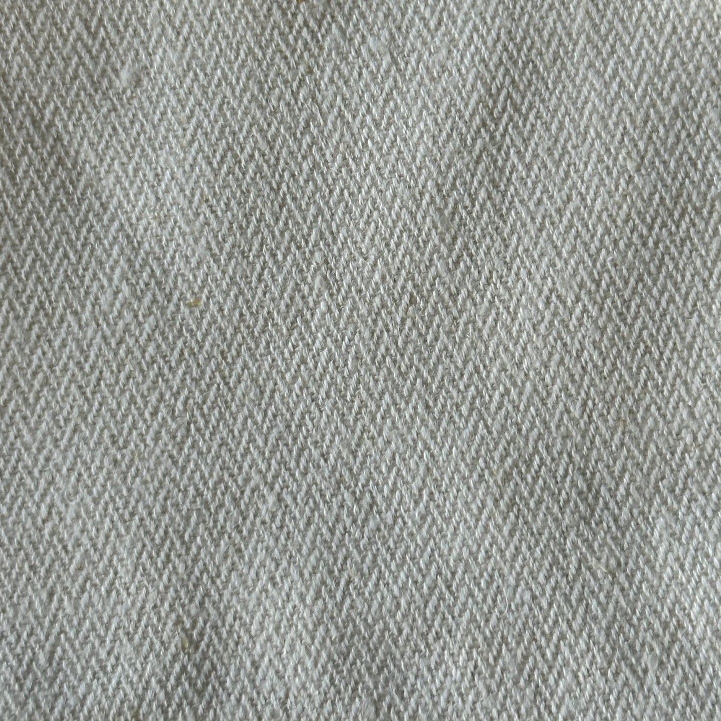 Close-up of a gray and beige herringbone fabric texture. The pattern is composed of small V-shaped weaving that alternates direction, creating a zigzag effect. Made from ethically handmade organic cotton twill, the fabric appears to have a soft and slightly fuzzy surface.