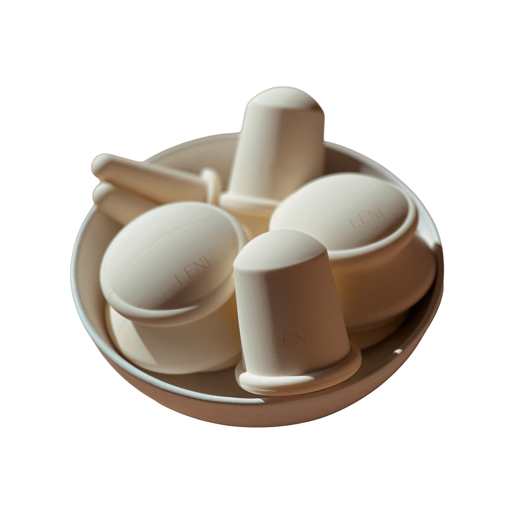 A white bowl contains various white, cup-like objects with handles from The Face & Body Cupping Set by LENI. They boast a sleek, modern design, and some are stamped with the brand name "LENI." Bathed in soft lighting, the items cast subtle shadows.