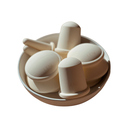 A white bowl contains various white, cup-like objects with handles from The Face & Body Cupping Set by LENI. They boast a sleek, modern design, and some are stamped with the brand name "LENI." Bathed in soft lighting, the items cast subtle shadows.