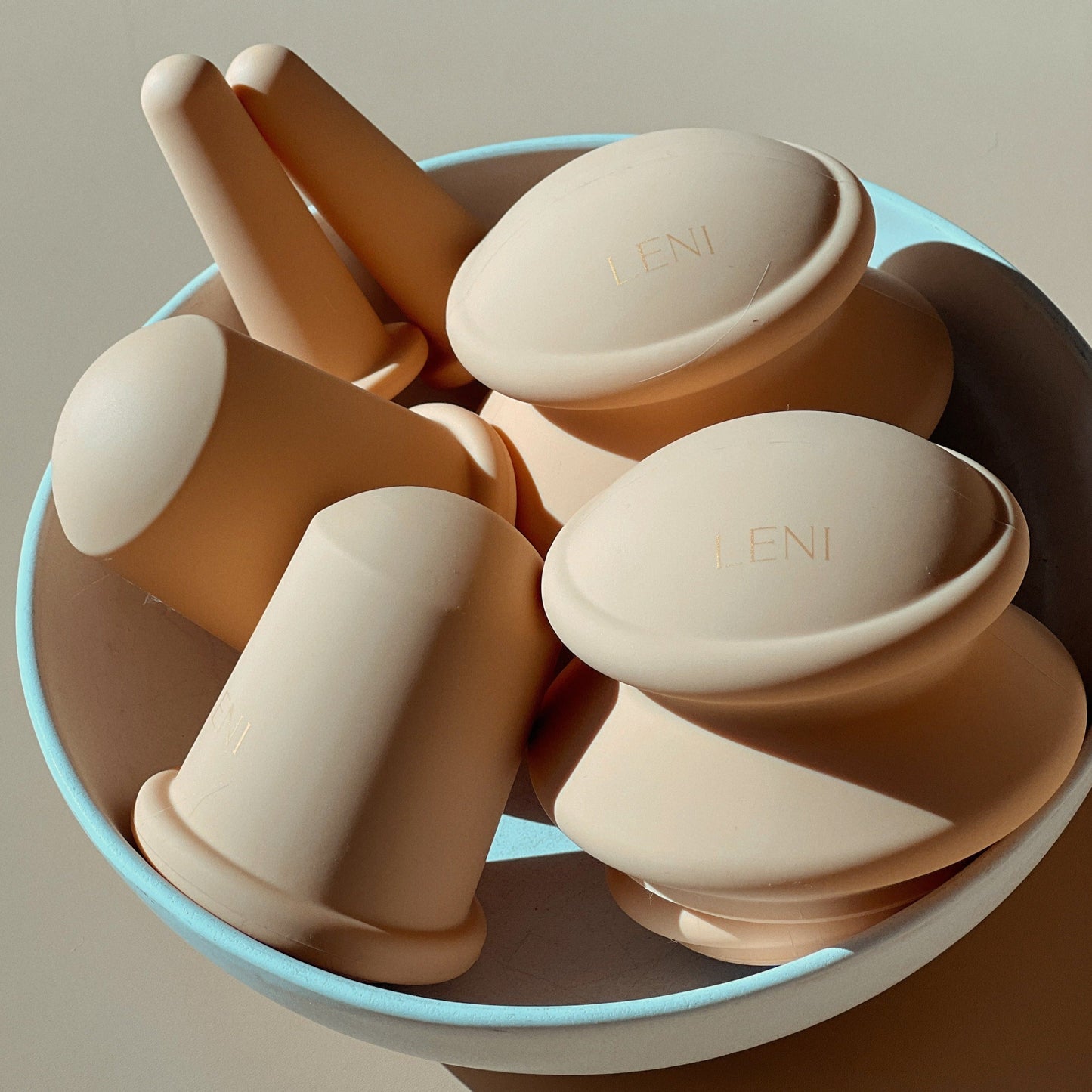 A bowl contains several beige silicone molds from The Face & Body Cupping Set by LENI, arranged under soft lighting, casting gentle shadows. Each mold has the word "LENI" embossed on its surface and showcases various shapes and textures designed for cupping therapy for the lymphatic system.