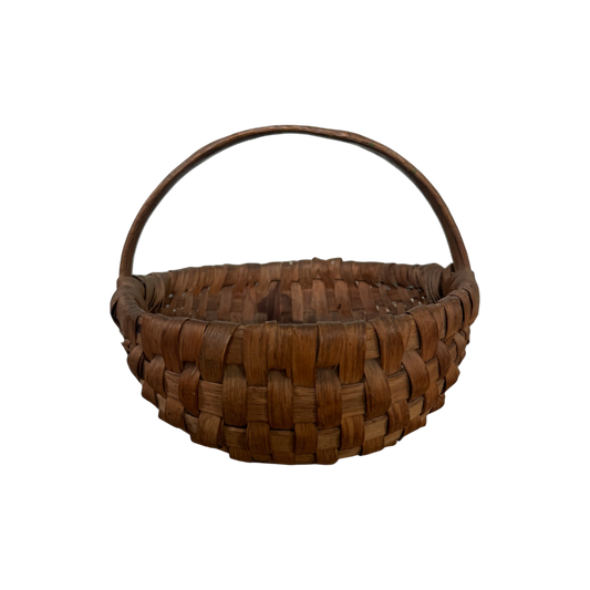 Antique Gather Basket I from Shoppe Maison, a round wicker basket featuring a curved handle, is displayed against a black background. Crafted from brown material, this woven basket exudes a rustic charm reminiscent of vintage goods.