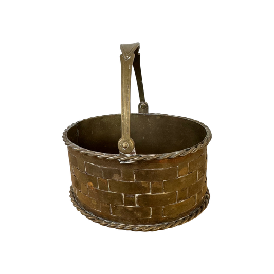 The Mini Brass Basket by Shoppe Maison is a rustic, round metal basket featuring a woven lattice pattern and a handle. This vintage piece is designed from brass and showcases twisted rope-like details along the base and top rim, all set against a solid black background.