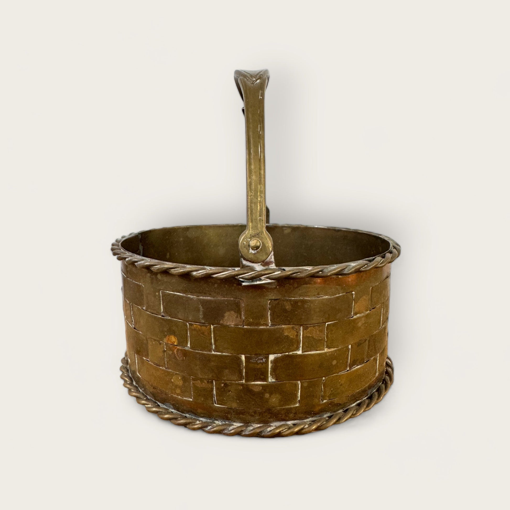 Introducing the Mini Brass Basket by Shoppe Maison: This cylindrical brass basket boasts a woven lattice detail and a rope-like trim accentuating both the top and bottom edges. It comes with a robust handle attached at the sides, allowing it to stand upright effortlessly. The plain, light-colored background highlights the vintage metallic texture of this captivating piece from Shoppe Maison's collection.