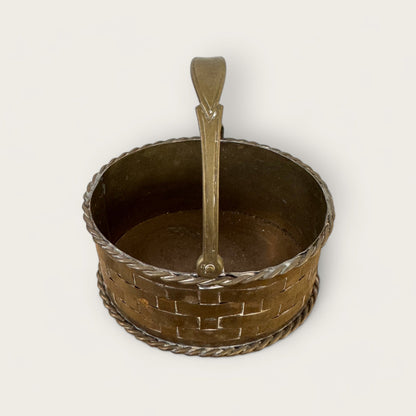 This is the Shoppe Maison Mini Brass Basket, a small and round piece adorned with a woven lattice detail and a braided edge. The basket features a sturdy, curved handle attached to the rim. Set against an off-white background, every intricate detail of this vintage good is beautifully highlighted.