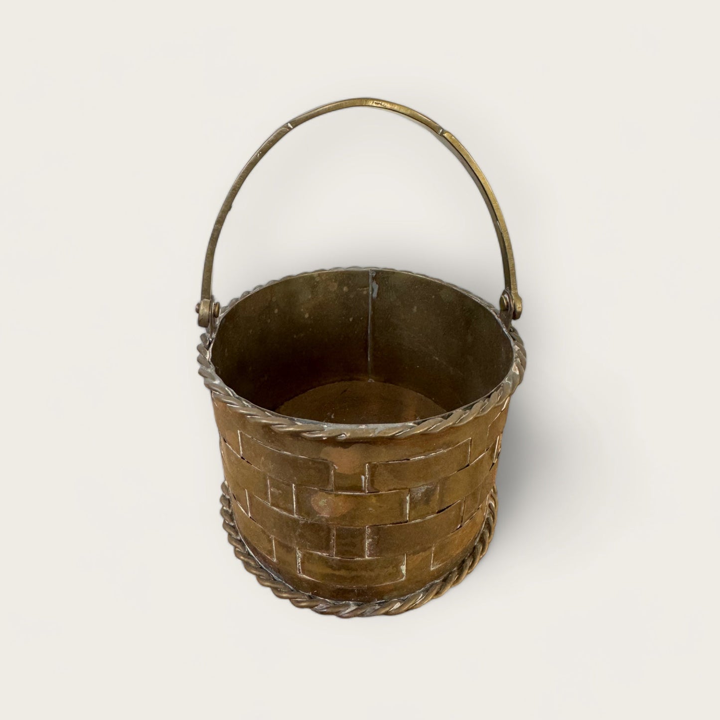 The Mini Brass Basket from Shoppe Maison is an antique, round metal bucket adorned with woven lattice details and decorative twisted metal trim along the top and bottom edges. It features a single curved handle attached on opposite sides, and its interior is plain without any additional design or markings—perfect for vintage goods enthusiasts.