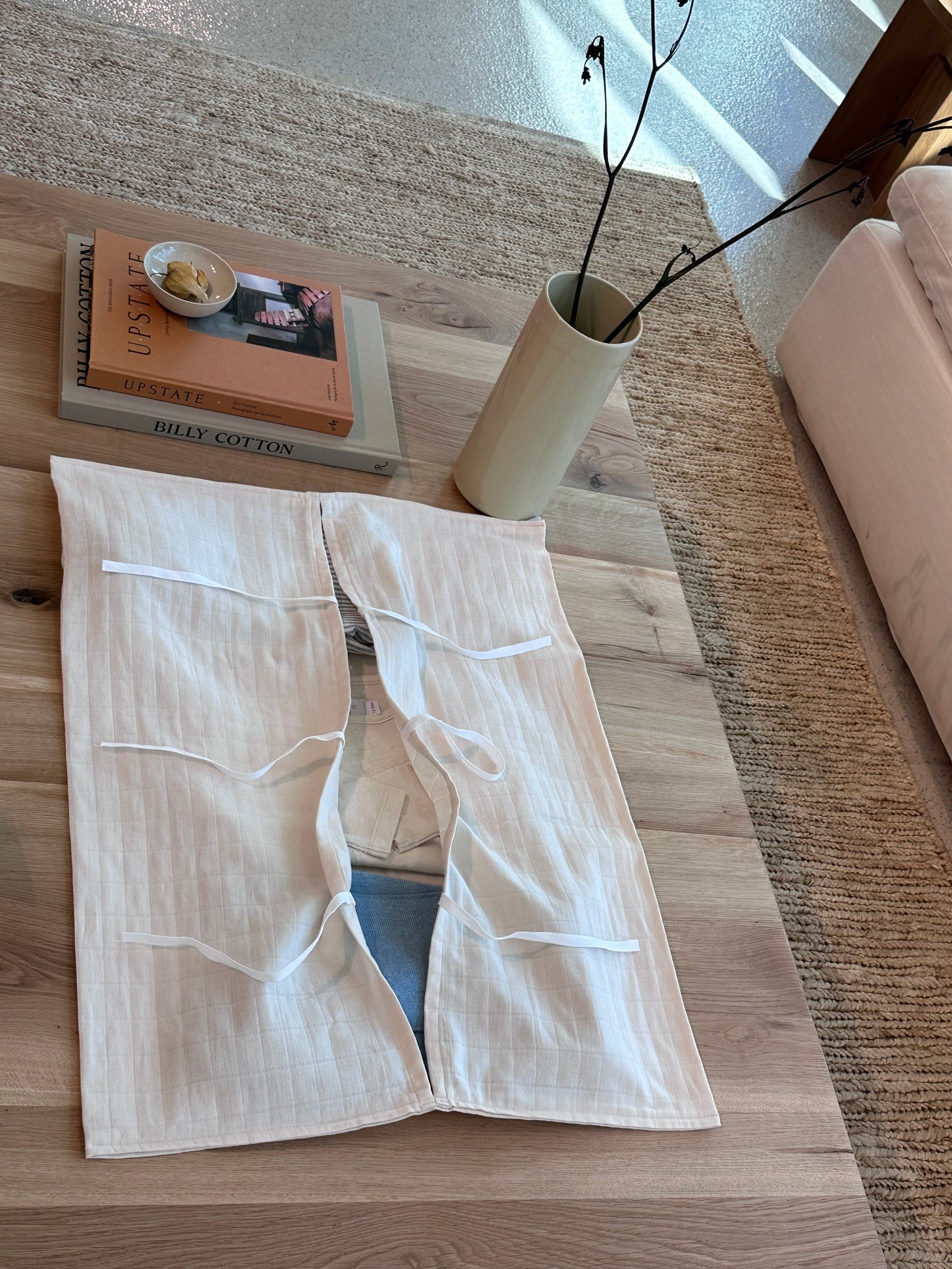 An Organic Garment Bag from cabane childrenswear in white linen with ties is partially open on a wooden coffee table, showcasing its organic cotton fabric. Nearby, there is a stack of books, a small bowl holding various items, and a tall vase containing thin branches. The coffee table rests on a beige woven rug beside a white sofa—making it ideal for storing children's clothing or as a lay-flat bag.