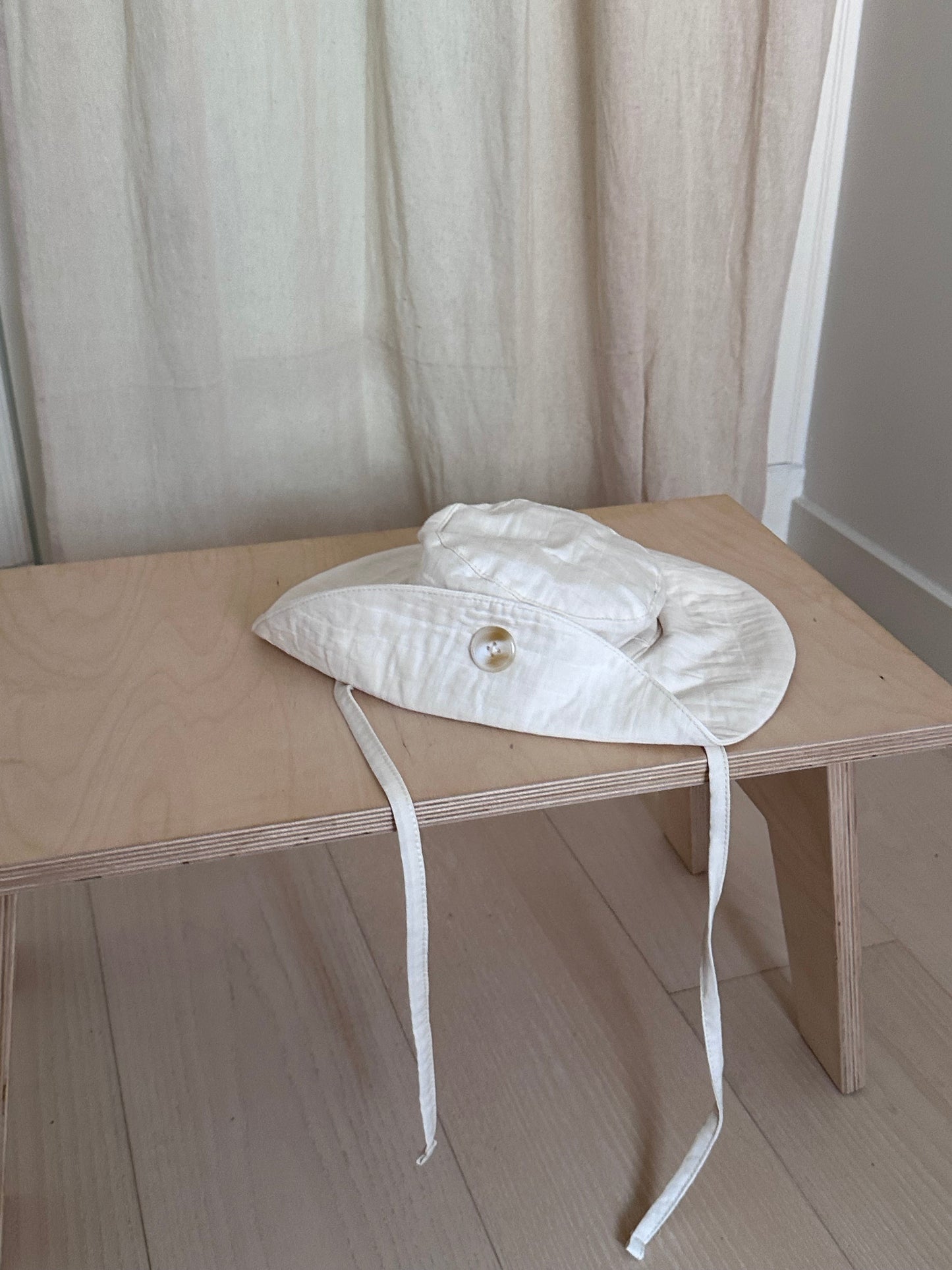 A white baby bonnet with ribbons is neatly placed on a light wooden bench. The bench stands on a light-colored floor, and a beige curtain hangs in the background, creating a soft and serene setting. Nearby, a "FRIENDS OF THE SUN" hat by cabane childrenswear, ethically handmade in Vancouver, adds an elegant touch to the scene.