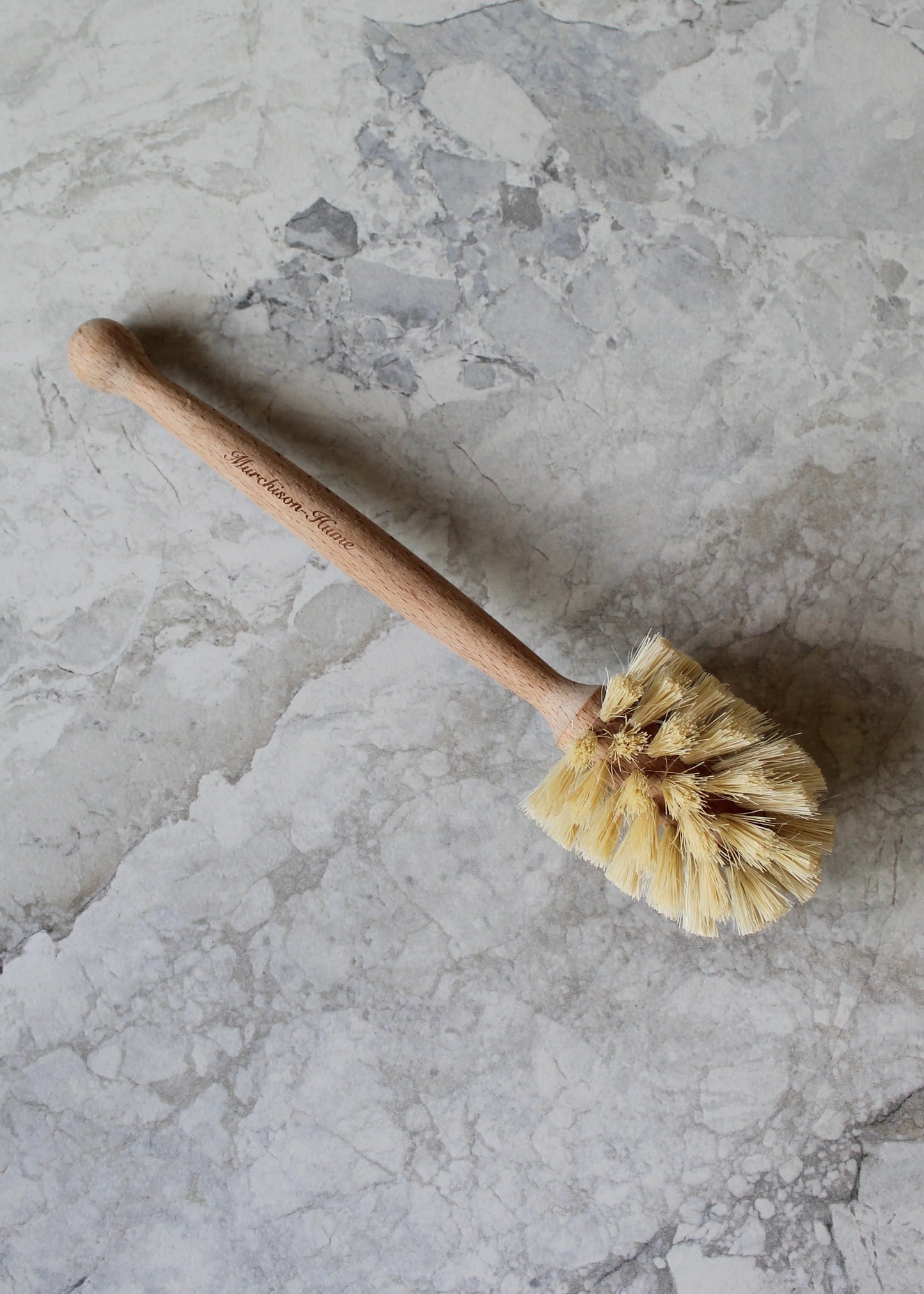 An eco-friendly dish brush with natural fiber bristles is placed on a gray marble surface. The handle, straight and smooth, features "Murchison-Hume" engraved along its length. This biodegradable All-Brush Bundle includes a brush head with stiff, densely packed bristles.