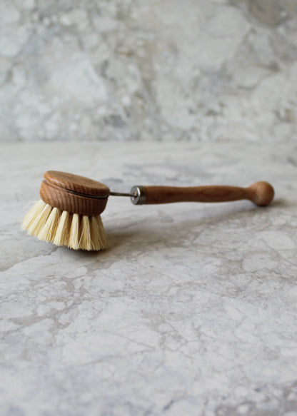 The All-Brush Bundle from Murchison-Hume, featuring a wooden dish brush with natural fiber bristles and a long handle, is placed on a light gray marble surface. The brush is positioned horizontally, showcasing its minimalist and biodegradable design.