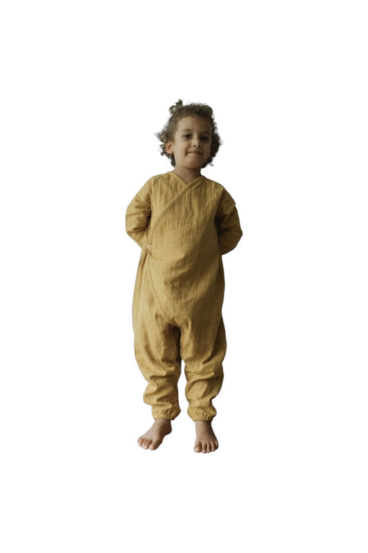 A young child with curly hair stands barefoot in a cozy mustard Après Wrap Palm jumpsuit made from organic cotton by cabane childrenswear, hands behind their back and smiling slightly against a plain white background.