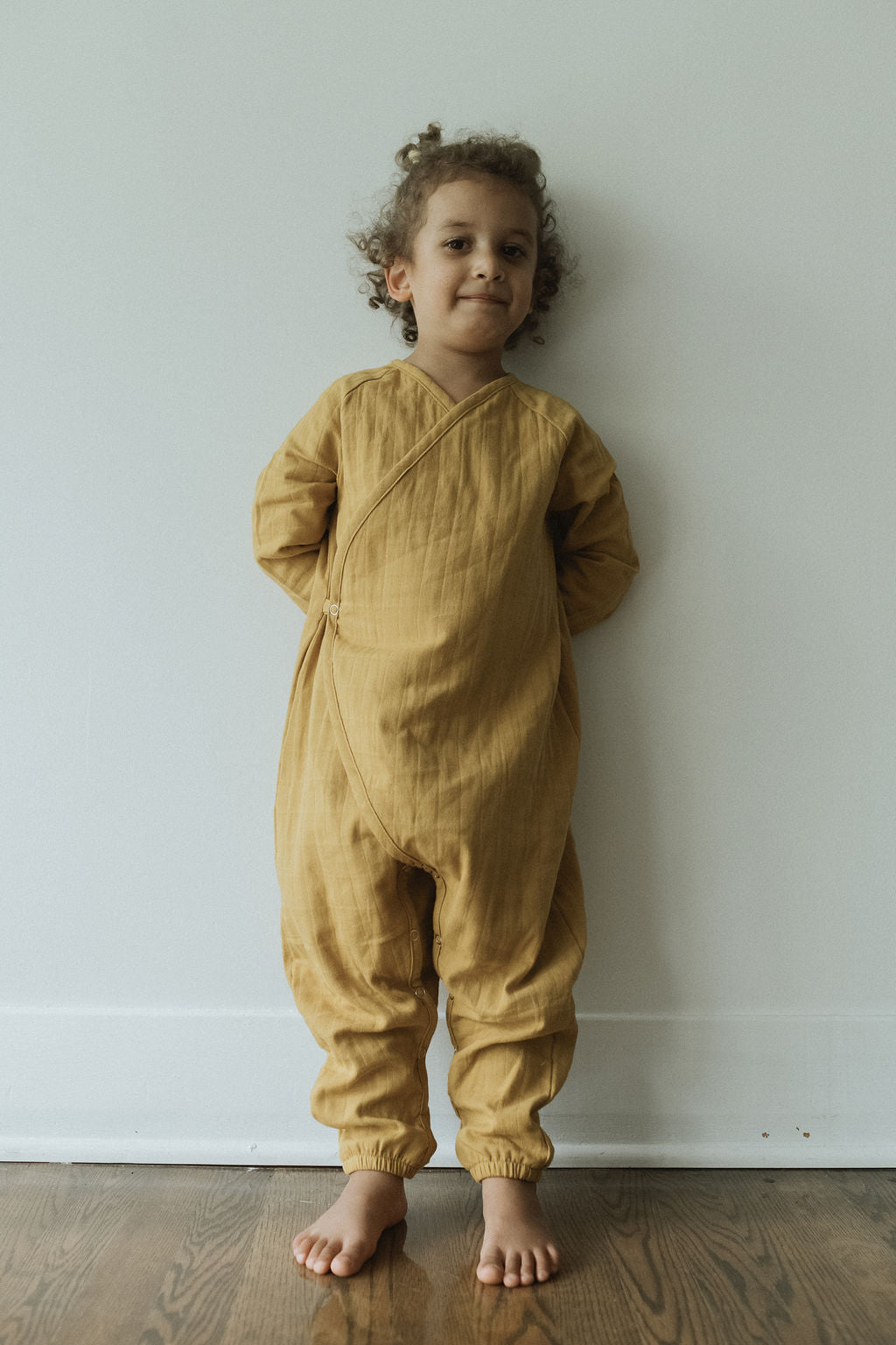 A child with curly hair stands barefoot on a wooden floor, wearing the APRÈS WRAP palm—a mustard-yellow, long-sleeved jumpsuit—from cabane childrenswear. Ethically handmade and crafted from breathable, organic cotton double gauze, they stand against a plain, light-colored wall with a slight smile.