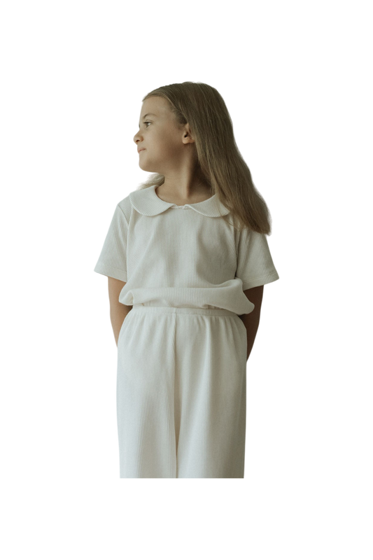 A young girl in a classic outfit stands against a plain background. She wears the pre-order SCHOOLHOUSE TEE waffle by cabane childrenswear, sporting long hair and a subtle gaze to her right, with her hands casually behind her back.