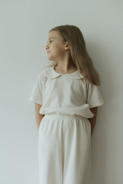 A young girl with long brown hair, standing against a white wall, looks to the side wearing the cabane childrenswear pre-order SCHOOLHOUSE TEE waffle. With hands behind her back, the scene is serene and minimalistic.