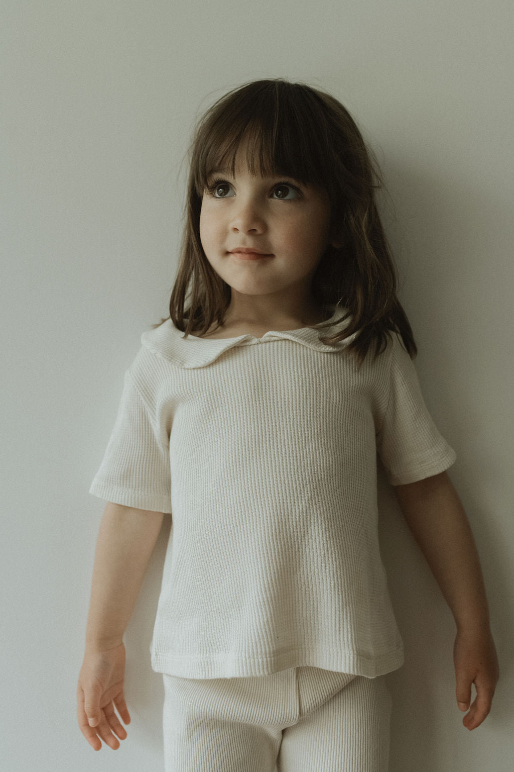 A young girl with long brown hair stands against a plain background, wearing the pre-order SCHOOLHOUSE TEE waffle by cabane childrenswear. She looks slightly upward with a small smile, embodying the brand's style, her hands resting by her sides.