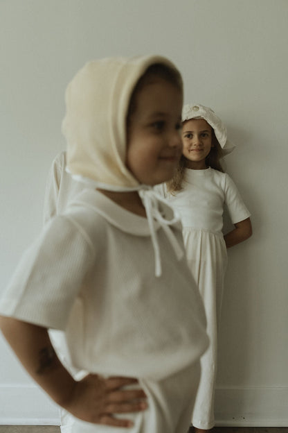 Two children in classic outfits, wearing the pre-order SCHOOLHOUSE TEE waffle by cabane childrenswear, stand against a plain wall. The child in front smiles warmly while the one behind playfully poses with hands on hips, creating a serene ambiance with timeless charm.