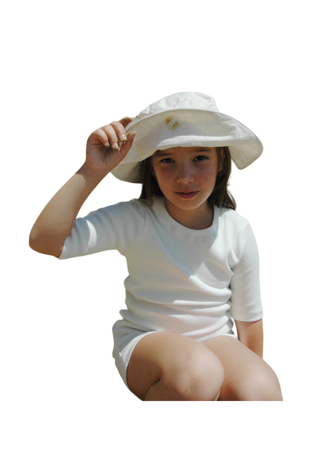 A young girl, dressed in a white outfit made of breathable summer material, sits cross-legged while smiling softly. She holds the brim of her "FRIENDS OF THE SUN" hat by cabane childrenswear with one hand, highlighting its ethically handmade quality from Vancouver. The background is plain black.