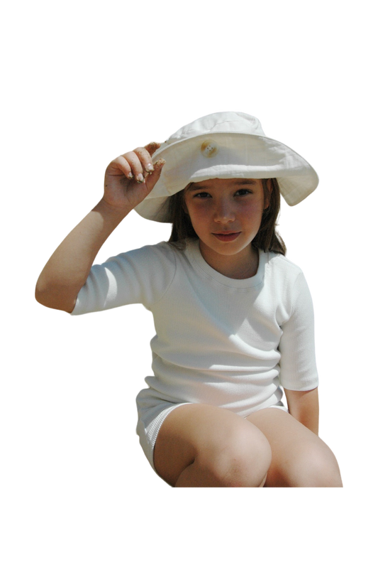 A young girl sits and looks at the camera, wearing a white outfit and the "Friends of the Sun" Hat by cabane childrenswear. She holds the brim with one hand, slightly tipping it up. The background has been removed, making the girl the sole focus of this ethically handmade summer wear image.