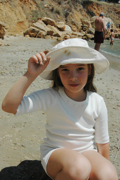 A young girl sits on a sandy beach near the water, adjusting her cabane childrenswear "FRIENDS OF THE SUN" hat and wearing a white outfit. In the background, people are enjoying the beach under a sunny sky, and rocky cliffs are visible along the shoreline.