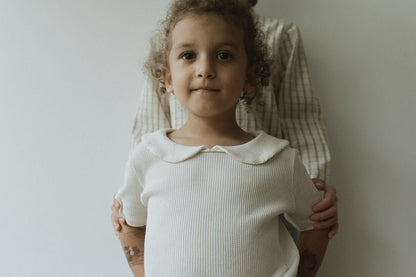 A young child, with curly hair in a pre-order SCHOOLHOUSE TEE waffle by cabane childrenswear, stands in front of an adult wearing a plaid shirt, as the adult's hands gently rest on the child's shoulders against a light-colored wall.