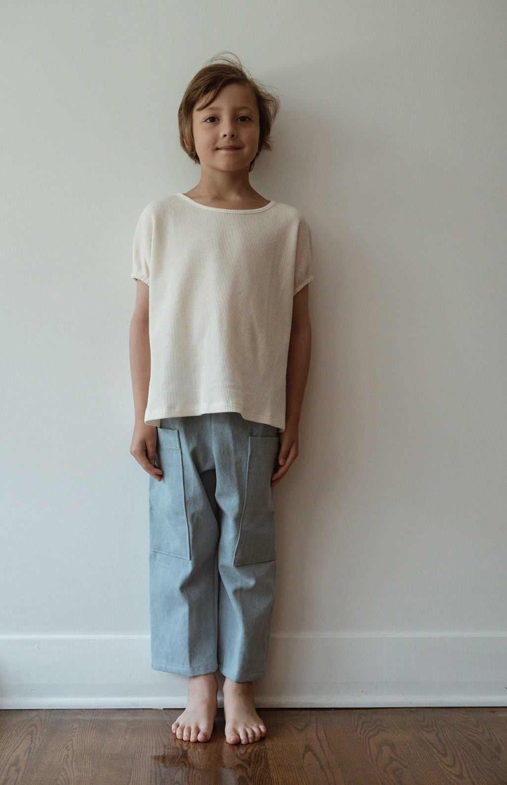 A child with short hair stands barefoot against a plain white wall, wearing the ethically handmade Pantalon D'Artiste in Denim by cabane childrenswear, paired with a loose white t-shirt and light blue shades. They're smiling softly at the camera. The wooden floor beneath them has a subtle sheen, reflecting the warmth of Calgary, Canada.