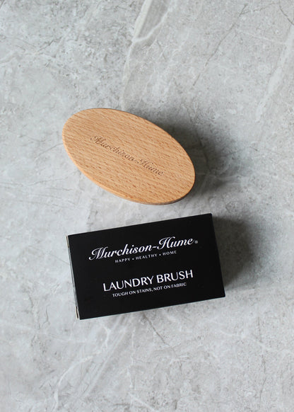 A wooden Murchison-Hume Compostable Laundry Brush with natural bristles and its black packaging, which reads "Murchison-Hume - Happy. Healthy. Home. Compostable Laundry Brush - Tough on stains, not on fabric," are placed on a light gray marble surface. Perfect for effective stain removal without damaging fabrics.