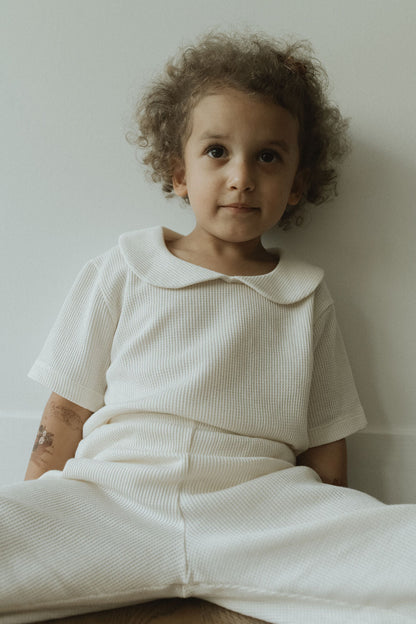 A child in a textured SCHOOLHOUSE TEE waffle from cabane childrenswear sits against a wall. The outfit's Peter Pan collar exudes classic style. They have curly hair and a calm expression while resting on the wooden floor with legs comfortably apart.