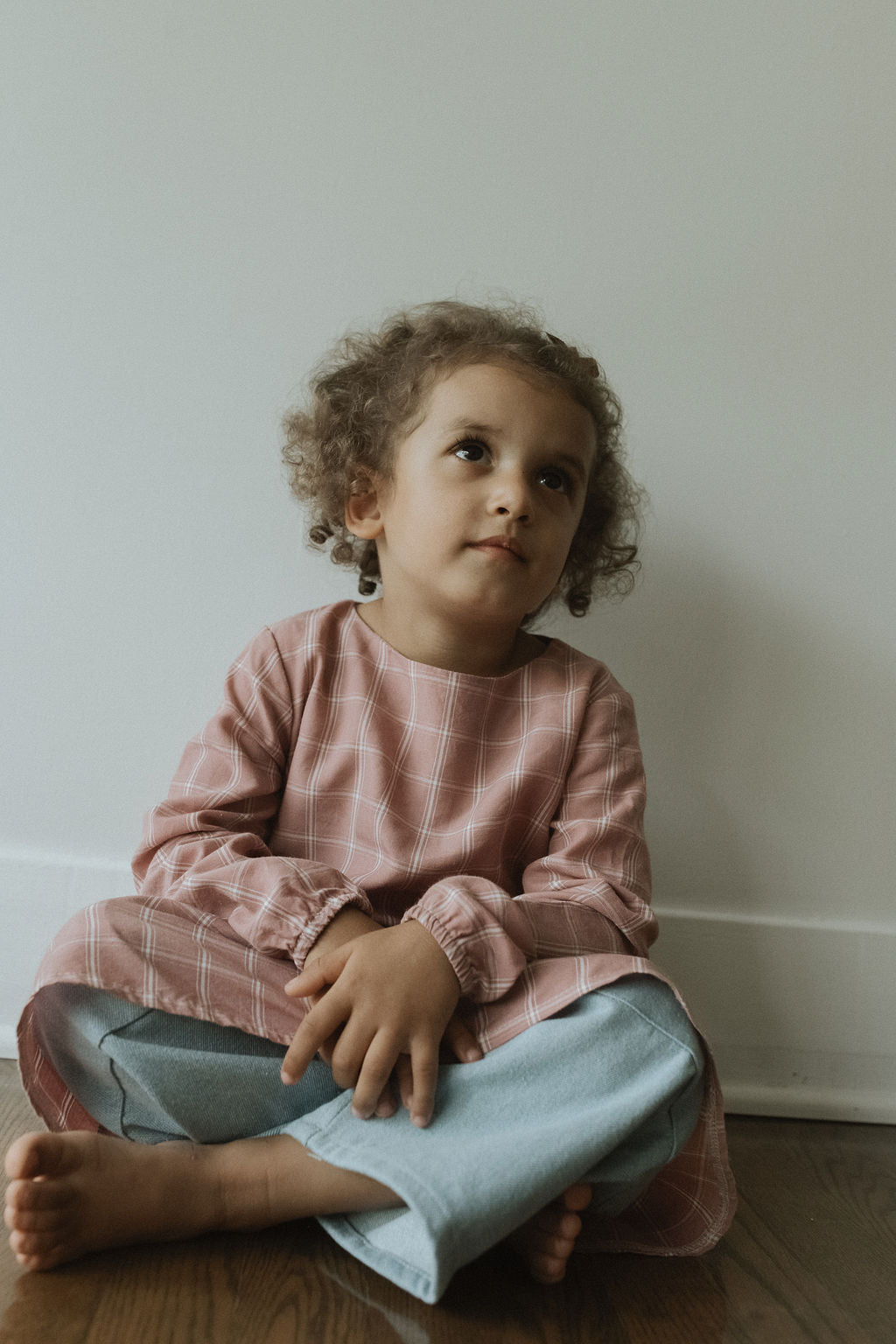 A young child with curly hair sits on a wooden floor against a white wall, gazing upward. Dressed in the limited edition raspberry plaid DAY DRESS by cabane childrenswear and light blue jeans, their hands rest gently in their lap.