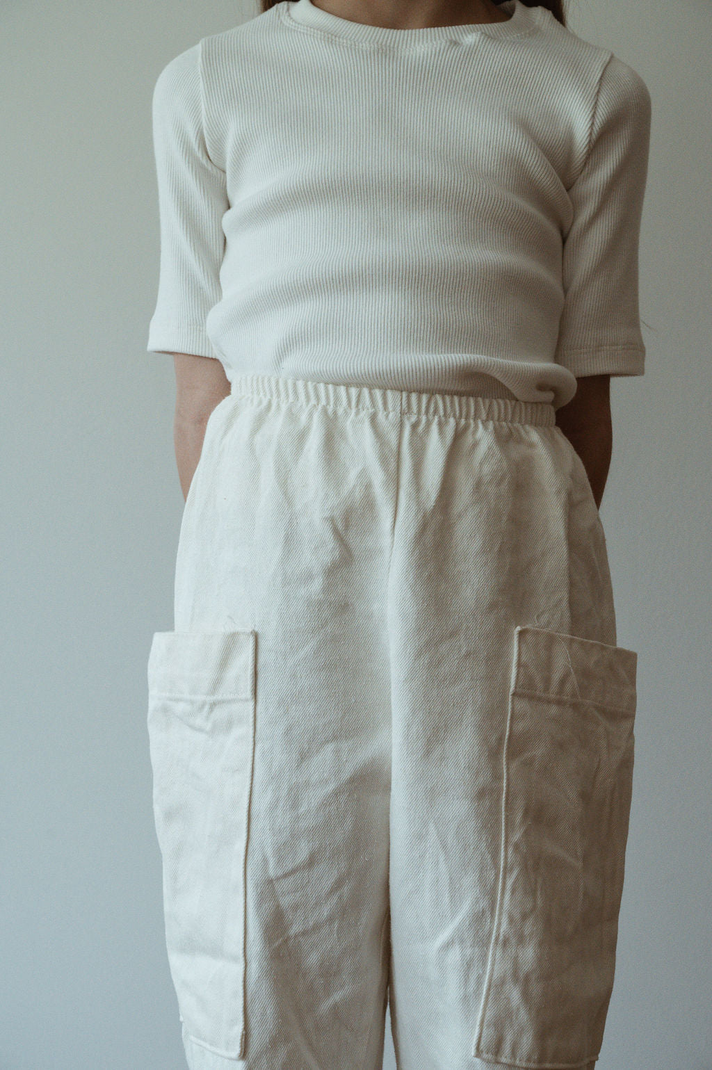 A person wearing a cream-colored ribbed short-sleeve shirt and cabane childrenswear's Pantalon D'Artiste in Hemp Twill stands against a plain background. The high-waisted pants feature large patch pockets on the front, and the person's face is not visible in the image.