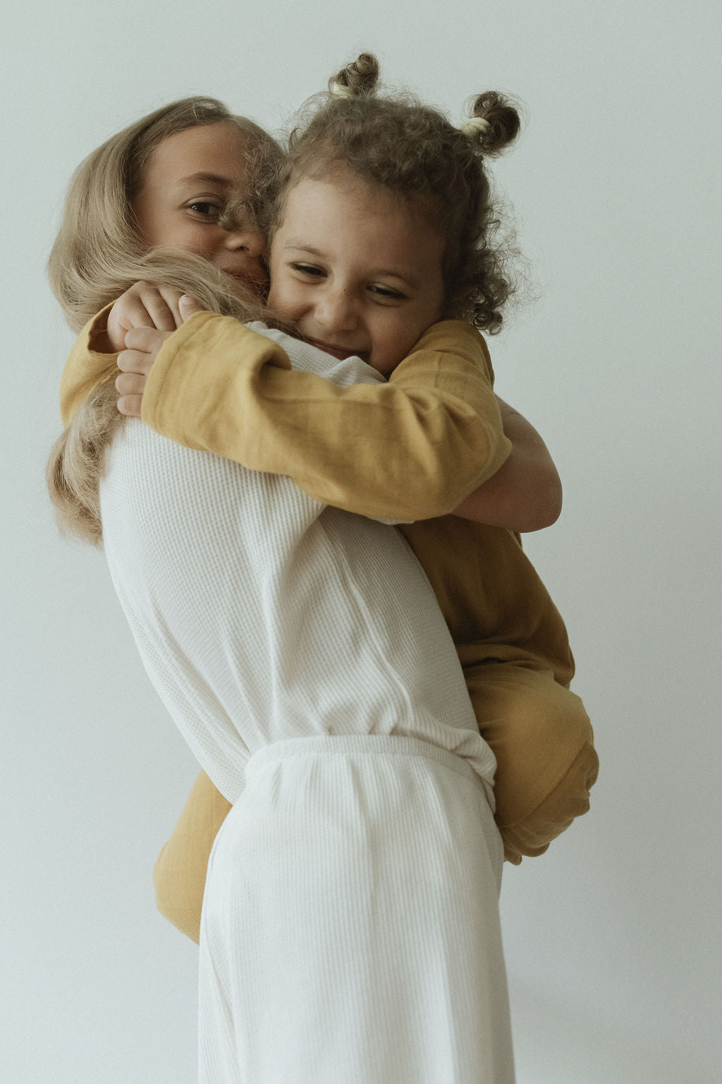 A person dressed in white lovingly holds a smiling child wearing the APRÈS WRAP palm outfit in a mustard color, crafted from ethically handmade, organic cotton by cabane childrenswear. The child's hair is styled in two small buns, and they are embracing the person closely, both appearing joyful and content.