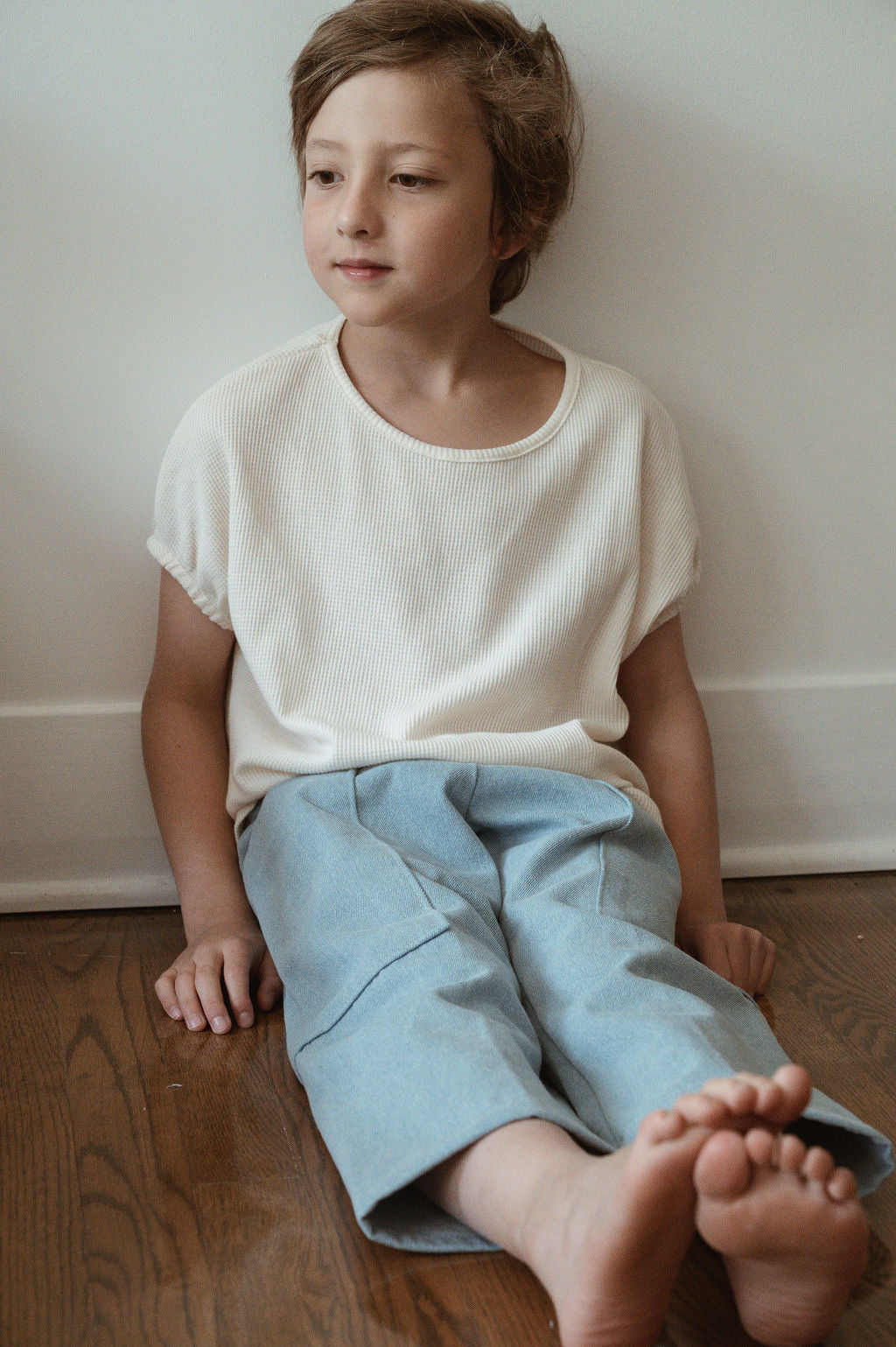 A young child with short hair sits on a wooden floor against a white wall, wearing an ethically handmade white shirt and Pantalon D'Artiste in Denim by cabane childrenswear. The child gazes slightly to the side, exuding a relaxed and thoughtful demeanor. The child's bare feet rest on the floor, enhancing the calming atmosphere in Calgary, Canada.