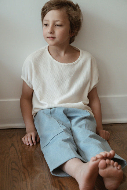 A young child with short hair sits on a wooden floor against a white wall, wearing an ethically handmade white shirt and Pantalon D'Artiste in Denim by cabane childrenswear. The child gazes slightly to the side, exuding a relaxed and thoughtful demeanor. The child's bare feet rest on the floor, enhancing the calming atmosphere in Calgary, Canada.