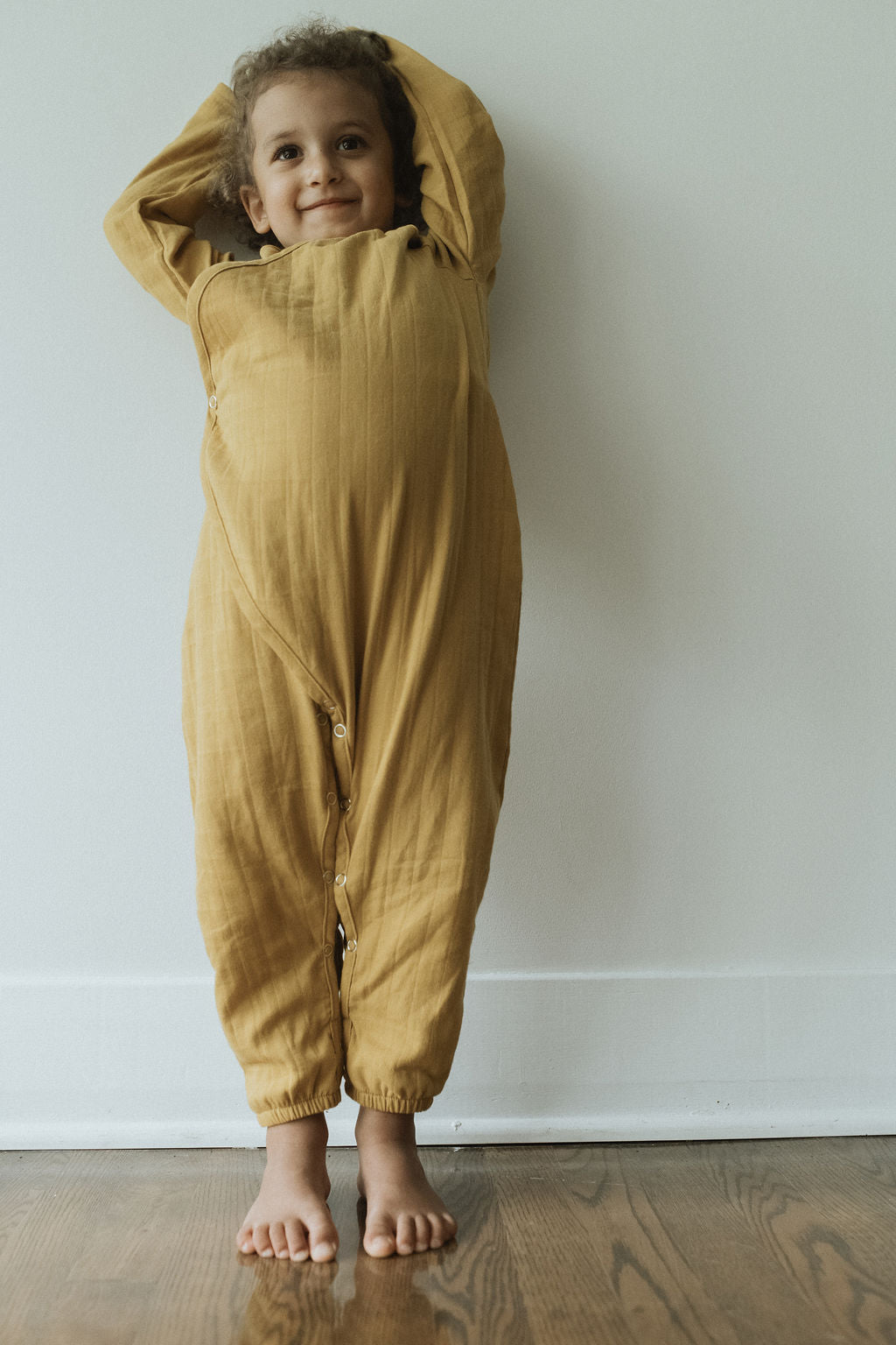 A child with curly hair smiles while standing barefoot against a wall, wearing the APRÈS WRAP palm by cabane childrenswear, made of breathable fabric. Their hands are raised above their head, and the wooden floor beneath them adds warmth to the scene.