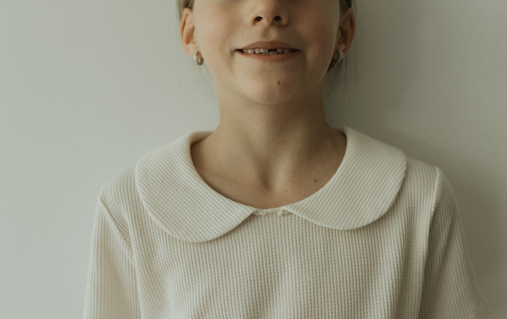 A young girl stands against a light-colored wall, smiling softly. She's wearing the pre-order SCHOOLHOUSE TEE waffle from cabane childrenswear, featuring a classic Peter Pan collar. Her look is complemented by small hoop earrings and her hair is neatly tied back, with the photo cropped at her shoulders.