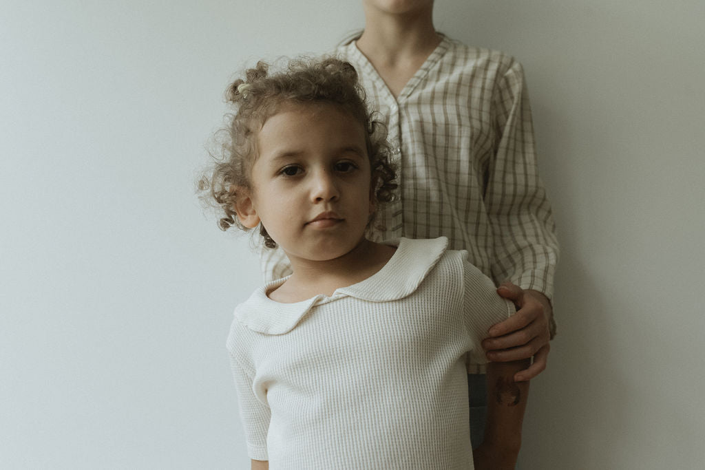 A young child with curly hair in a classic pre-order SCHOOLHOUSE TEE waffle stands before an older child in a plaid shirt, showcasing timeless style. The older's gentle arm rests on the younger one's shoulder, embodying cabane childrenswear elegance against a pristine white backdrop.