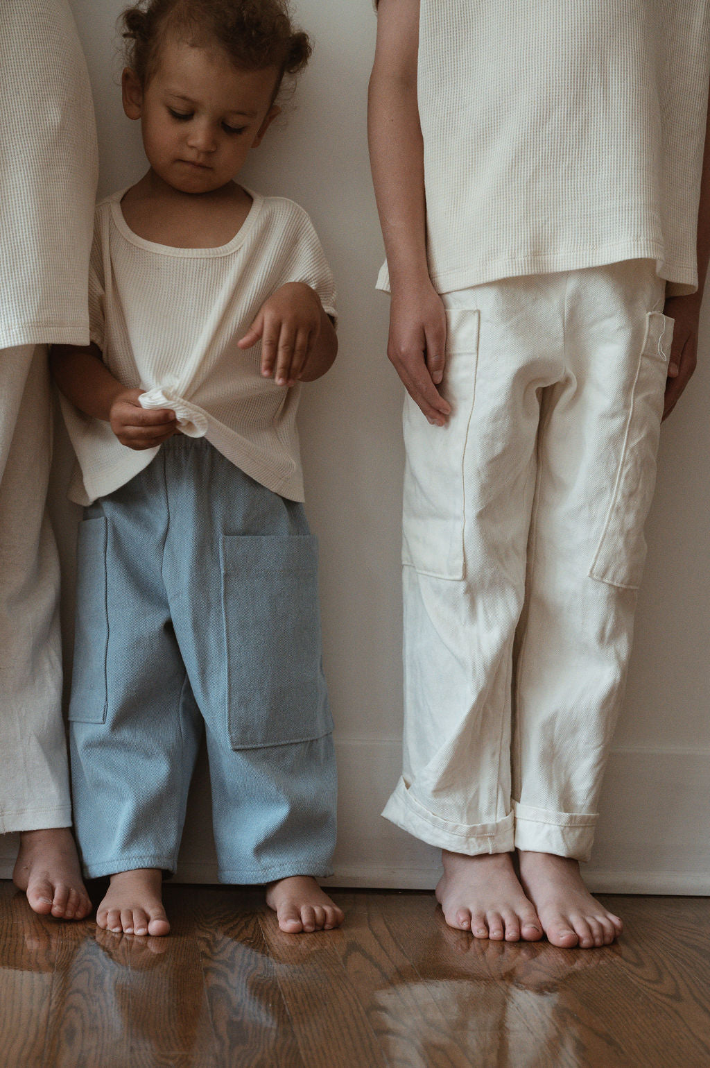 Two children stand barefoot on a wooden floor against a white wall. The child on the left wears a cream top and cabane childrenswear's Pantalon D'Artiste in Denim, gazing downward. To the right, another child is dressed in cream-colored clothing. Both children are partially out of frame, and their outfits are ethically handmade in Calgary, Canada.