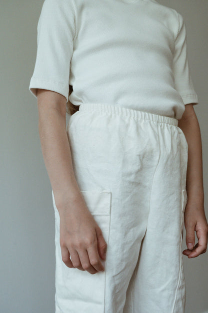 A person wearing a short-sleeved ribbed white shirt tucked into the Pantalon D'Artiste in Hemp Twill by cabane childrenswear, which features a high-waisted, loose-fitting design with side pockets, stands against a plain background.