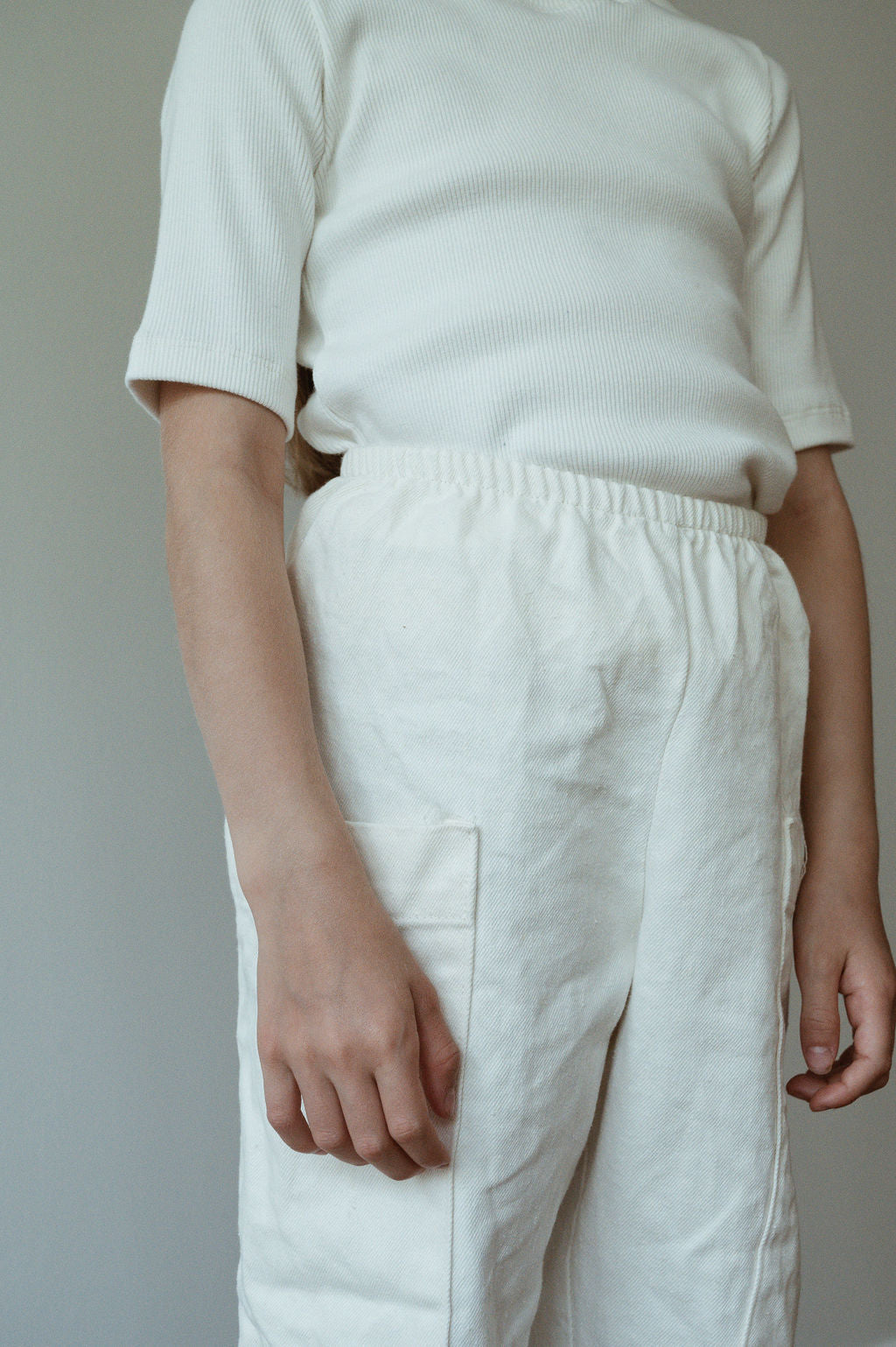 A person styled in cabane childrenswear's ethically handmade Pantalon D'Artiste in Hemp Twill, cream-colored for texture and simplicity, stands against a neutral backdrop wearing an off-white ribbed short-sleeve shirt. This look highlights a dedication to craftsmanship.