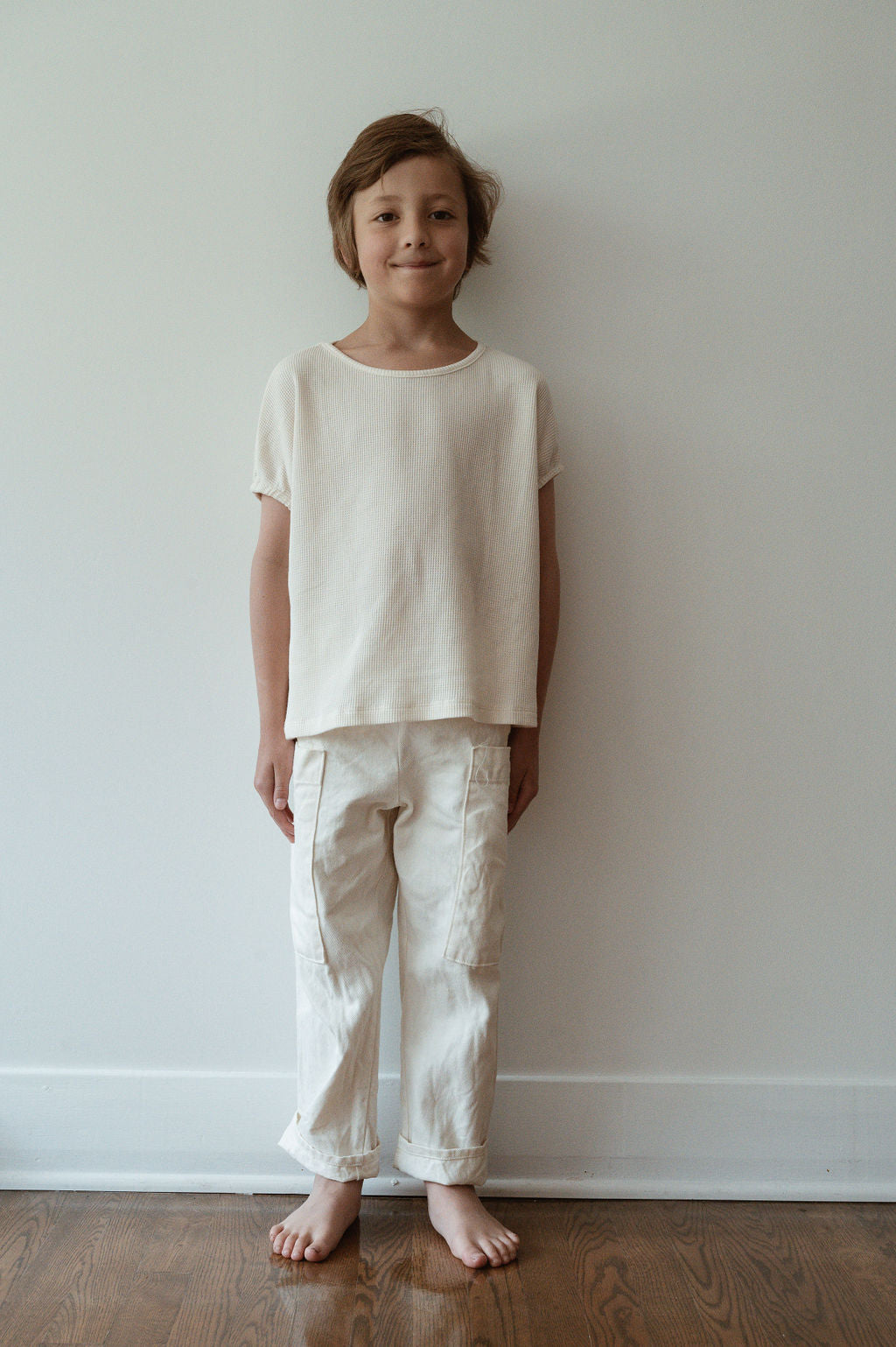 A young child, dressed in cabane childrenswear's Pantalon D'Artiste in Hemp Twill, stands barefoot on a wooden floor against a plain white wall. They smile gently at the camera, exuding both innocence and eco-friendly charm.