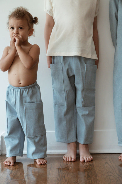 Two young children stand indoors on a wooden floor. The child on the left, shirtless and wearing cabane childrenswear's Pantalon D'Artiste in Denim, bites their fingers. The child on the right wears a white shirt with similar ethically handmade pants from the same brand. Both are barefoot, facing forward in a cozy room in Calgary, Canada.