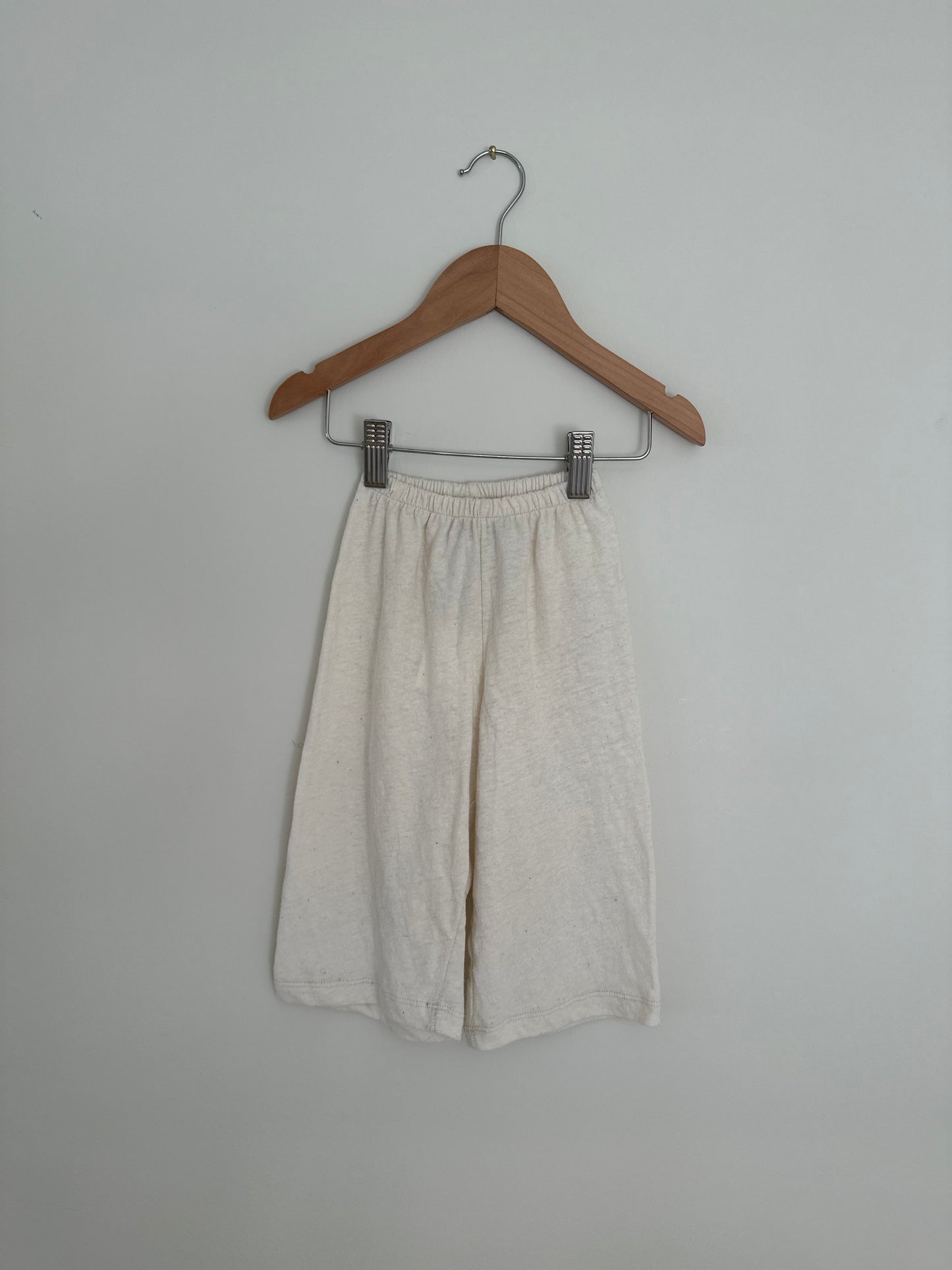 A pair of "Resort Pant in Hemp" by cabane childrenswear hangs on a wooden hanger against a plain white background. These ethically handmade pants feature an elastic waistband and a slightly wrinkled texture. The simple hanger has a hook at the top for hanging, evoking the serene feel of Calgary’s natural beauty.