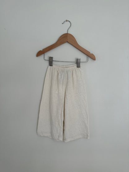 A pair of "Resort Pant in Hemp" by cabane childrenswear hangs on a wooden hanger against a plain white background. These ethically handmade pants feature an elastic waistband and a slightly wrinkled texture. The simple hanger has a hook at the top for hanging, evoking the serene feel of Calgary’s natural beauty.