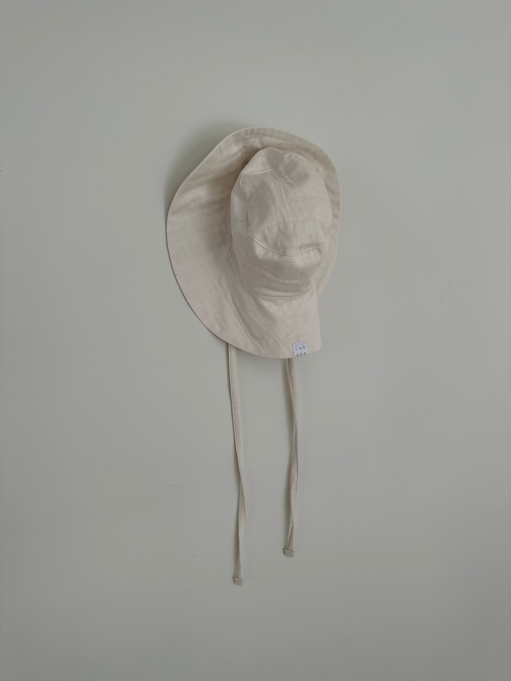 A "FRIENDS OF THE SUN" hat by cabane childrenswear, beige and classic with a wide brim and attached adjustable chin straps, hangs on a white wall. Ethically handmade in Vancouver, the hat is crafted from a light, breathable summer material that is perfect for sun protection.