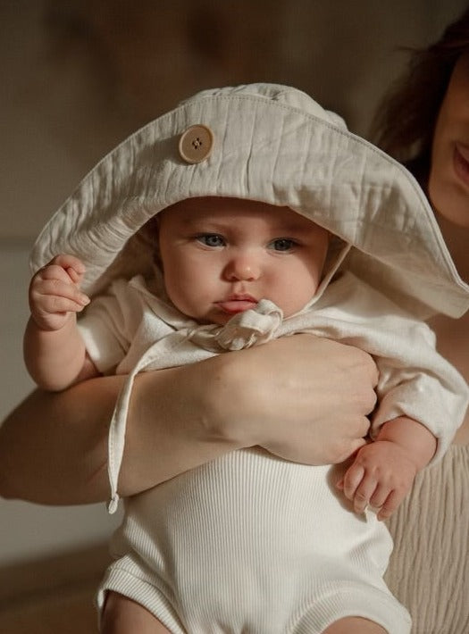 A baby wearing a cabane childrenswear "FRIENDS OF THE SUN" hat is being held by someone partially visible in the background. The baby has a serious expression and is dressed in a white outfit that matches the hat. The background is softly lit, creating a warm atmosphere.