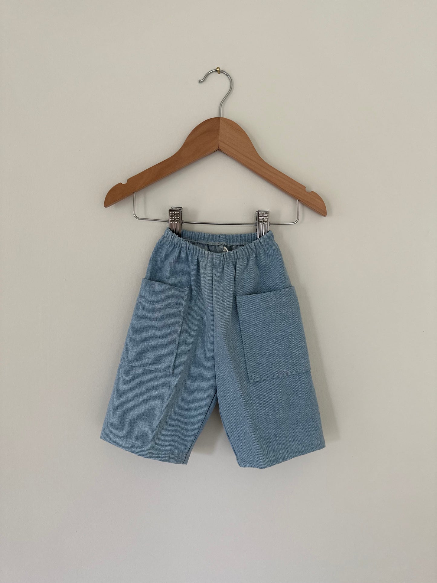 Photo of the "Pantalon D'Artiste in Denim" by cabane childrenswear, ethically handmade in Calgary, Canada. These light blue children's shorts are displayed on a wooden hanger, featuring an elastic waistband and two large front pockets, set against a plain off-white background.