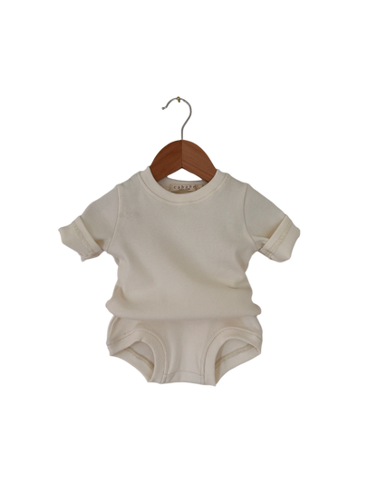 A cream-colored baby onesie with short sleeves and folded cuffs is hanging on a wooden hanger against a black background. The onesie, part of the CLASSIC SET baby + kid from cabane childrenswear, features a simple ribbed texture.