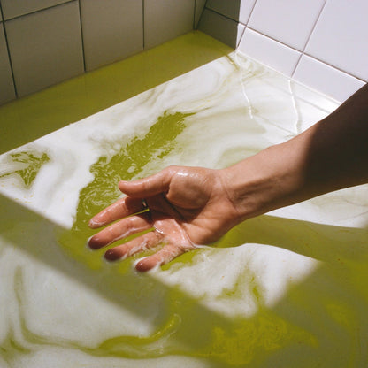 A person's hand, with water droplets on their fingers, dips into an Inheal® Probiotic Saffron Milk Bath by THE FULLEST, filled with yellow and white swirling water. Surrounded by white tiled walls, the lighting creates a reflective, serene atmosphere perfect for skin hydration.